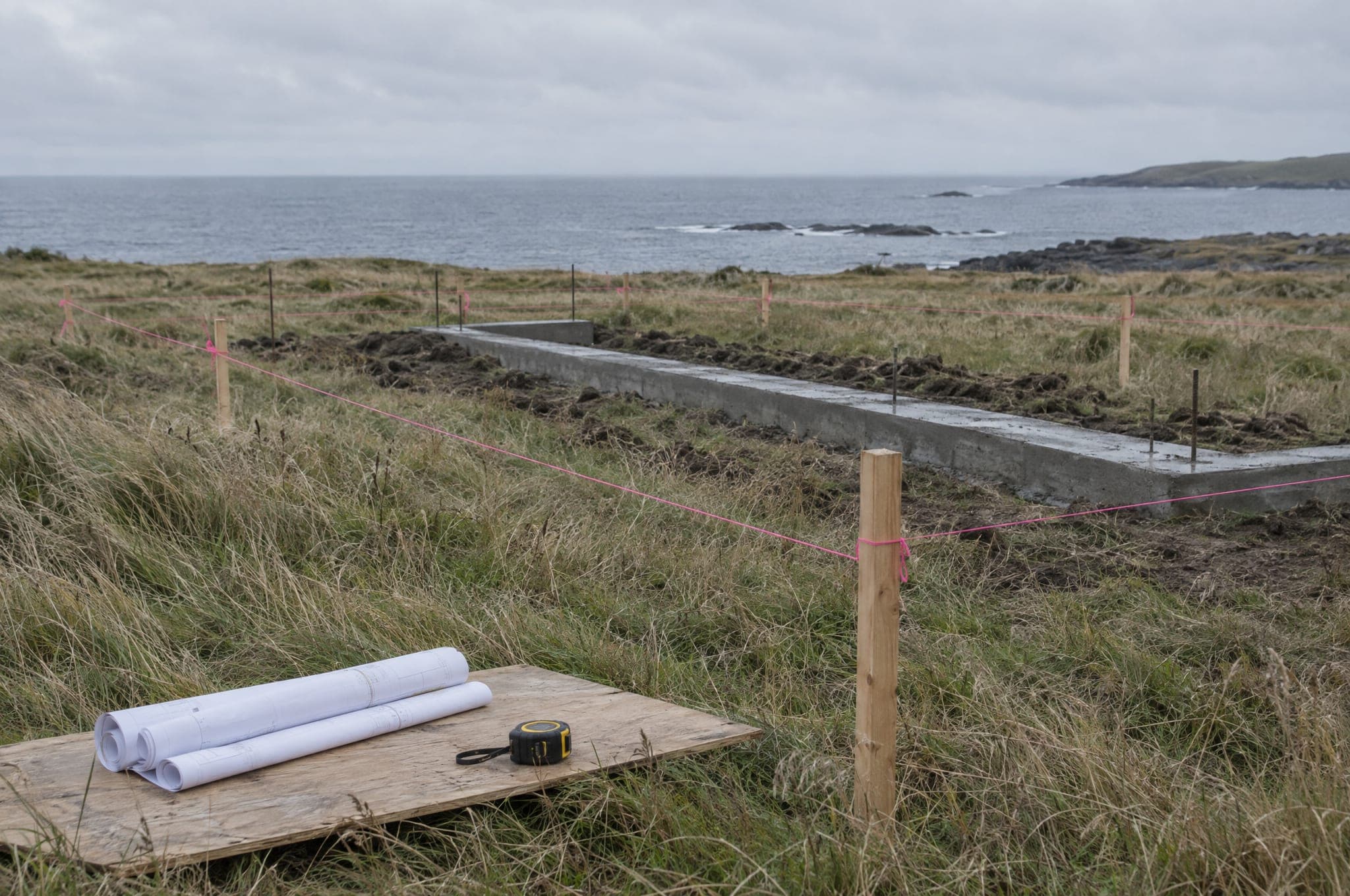 A building site before construction: survey stakes and string lines in long grass, rolled drawings, a fresh strip foundation