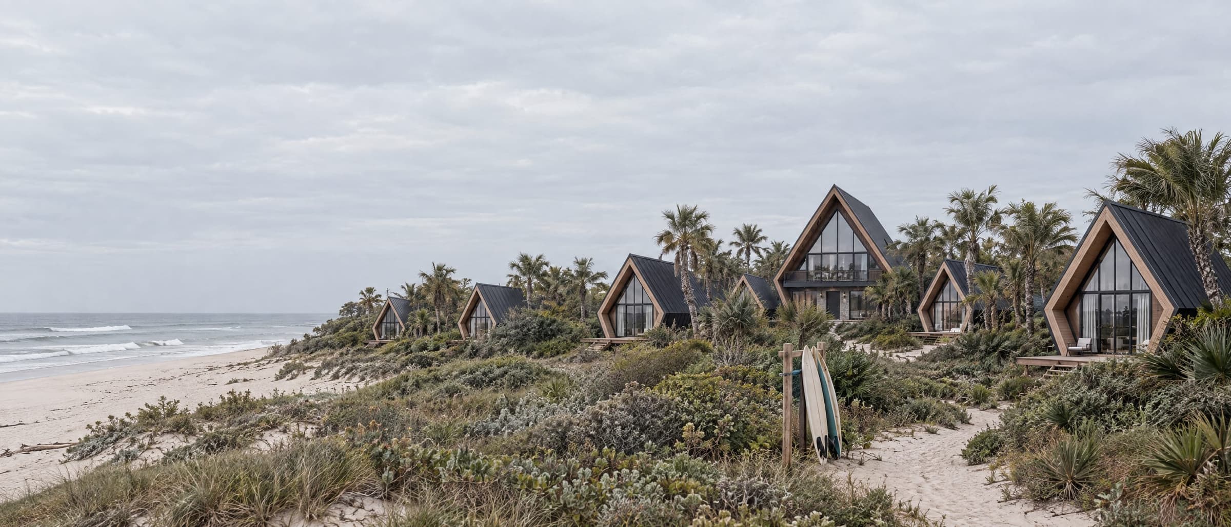 A surf camp set back from the beach: Studio cabins among dune scrub and palms behind the sand, surfboards on a rack, grey ocean beyond