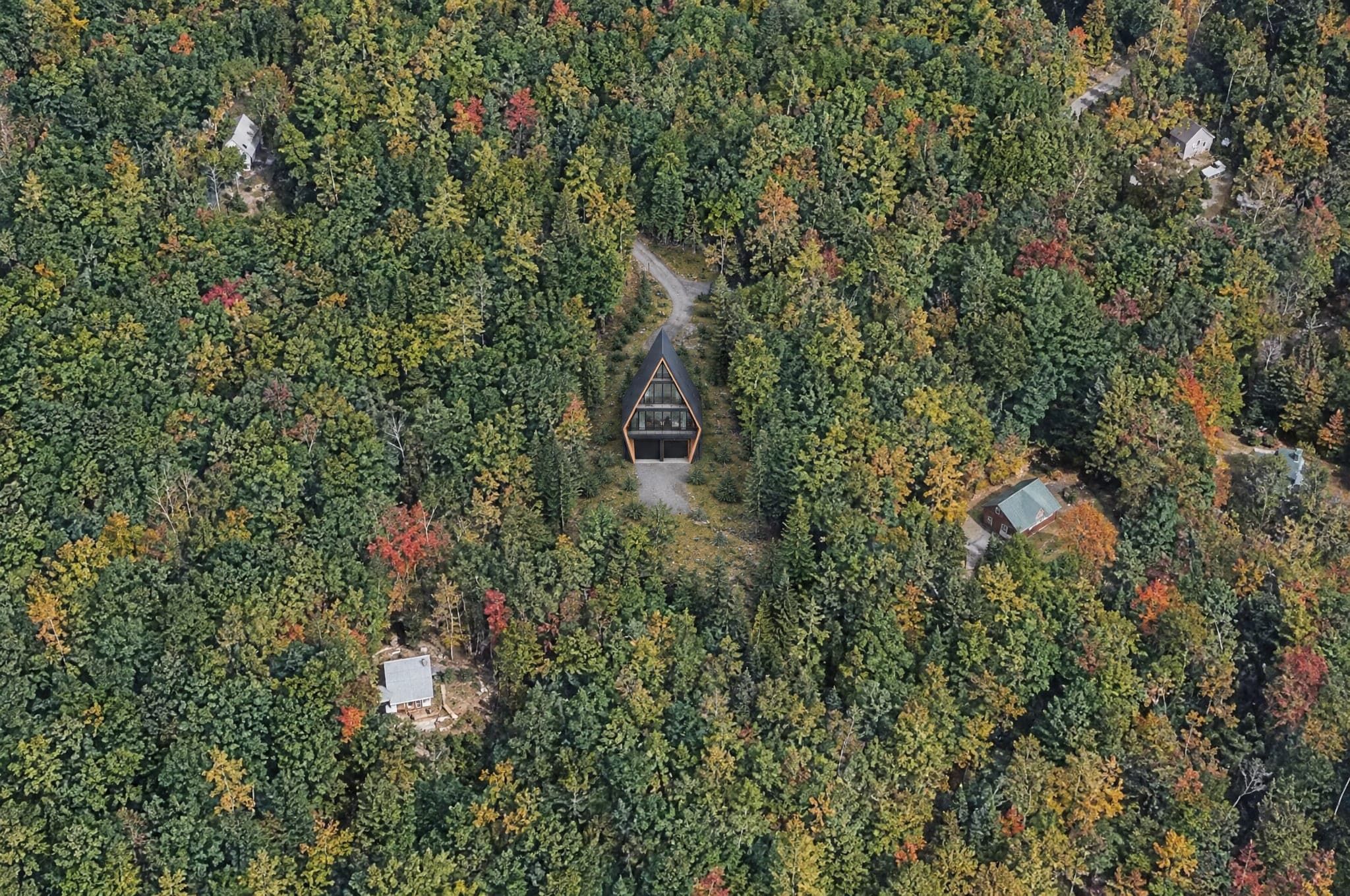 Aerial view of a wooded hillside lot in Waterville Estates, Campton, New Hampshire, with an AFRME Four A-frame placed among autumn trees beside the association road