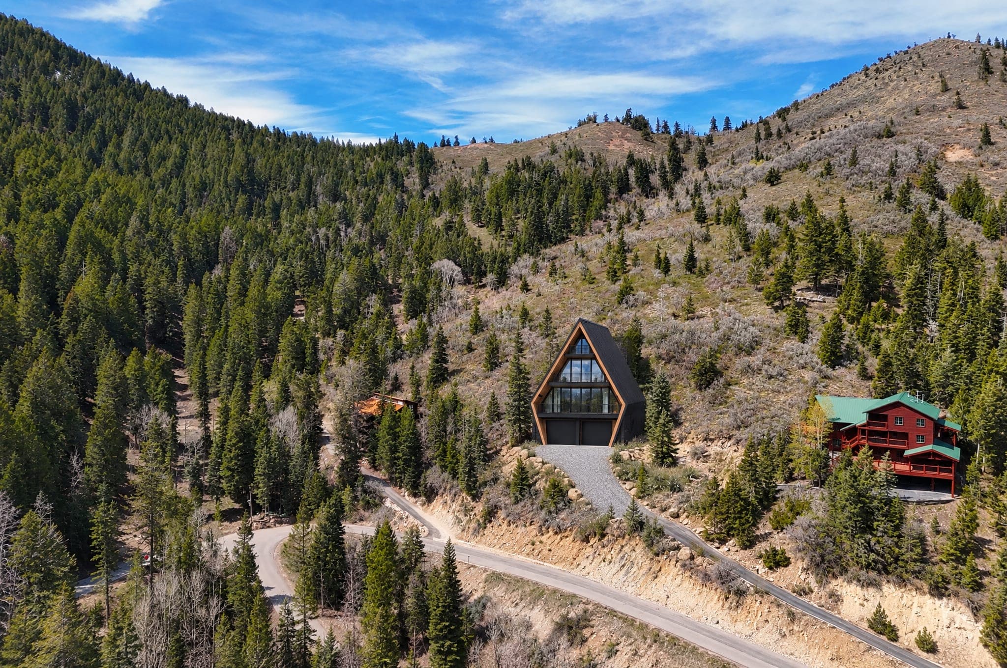 Drone view of a south-facing mountain lot in Summit Park, Utah, with an AFRME Four A-frame standing above Matterhorn Drive among firs and sagebrush, the ridge rising behind